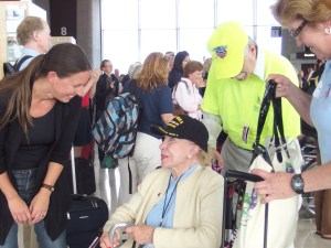 An older woman in a wheelchair in an airport terminal.  She wears a ballcap that says World War II veteran.  She is beautiful and is wearing elegant make up, nail polish and jewelry, as well as a blue polo shirt and white sweater.  To the left of the photo, a younger woman leans in, smiling, to speak to the woman in the wheelchair, while a man in a bright yellow shirt and hat stands behind the wheelchair.  In the back ground, a crowd of people look toward an airport gate, clapping.  