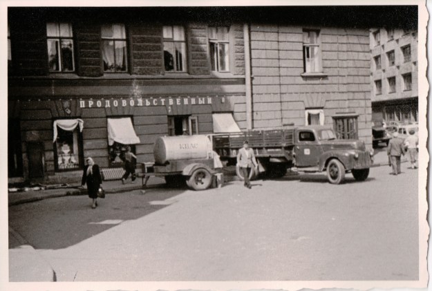 {Black and white photo of an old time pick up truck towing a small tank of liquid, parked in front of a building.}