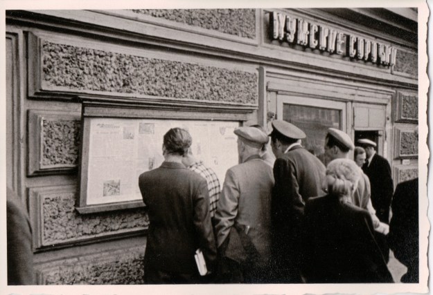 {Black and white photo of men gathered in front of a newspaper that is posted on the exterior wall of a building.}