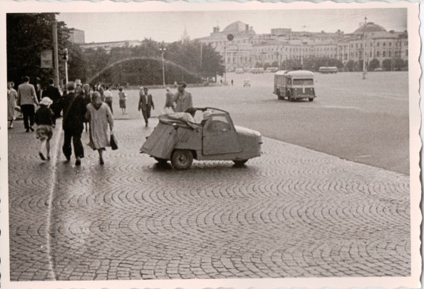 {Black and white photo of a cobblestone street and sidewalk.  In the foreground is a very small three-wheeled vehicle, suitable for at most one person, open on top and looking almost home-made out of pieces of welded steel.  In the background are pedestrians and in the far background, indistinct buildings.}