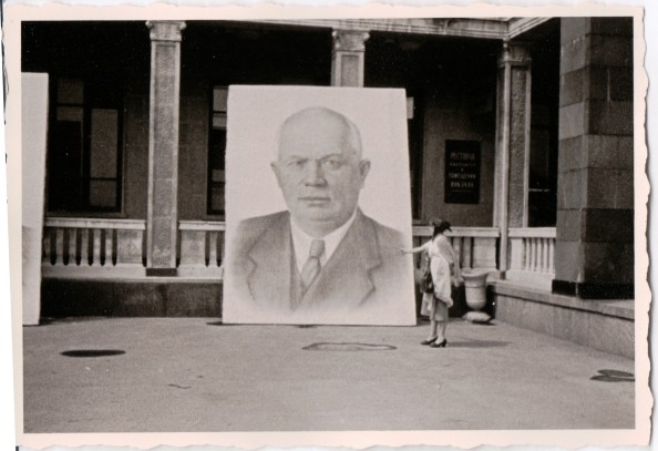 {Black and white photo of a building with a giant portrait of Nikita Khruschev leaning against the columns in the front of the building.  The portrait is over twice the height of a man standing near it.}