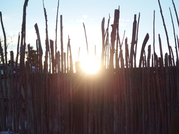 {Image:  fence made of vertical pieces of natural wood of varying heights.  In the bottom half of the photo, the wood slats are backed by a solid piece of wood.  In the top half of the photo, there is nothing behind the vertical slats, and the sun peeks through between two of the slats.}