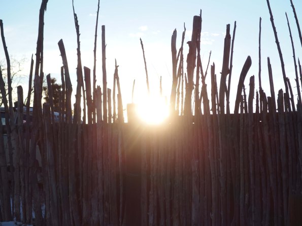 {Image:  fence made of vertical pieces of natural wood of varying heights.  In the bottom half of the photo, the wood slats are backed by a solid piece of wood.  In the top half of the photo, there is nothing behind the vertical slats, and the sun peeks through between two of the slats.}