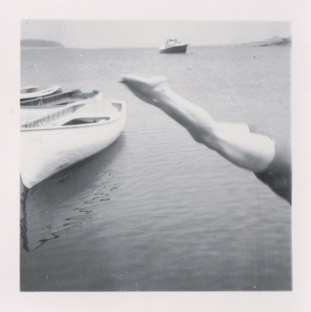 {Image: black and white photo of a person diving into the water, but all we see is the person's legs, perfectly straight, angled from their toes at the center of the photo to the bottom right where their torso disappears off the edge of the photo. To the left are several canoes, and in the background, a boat.}