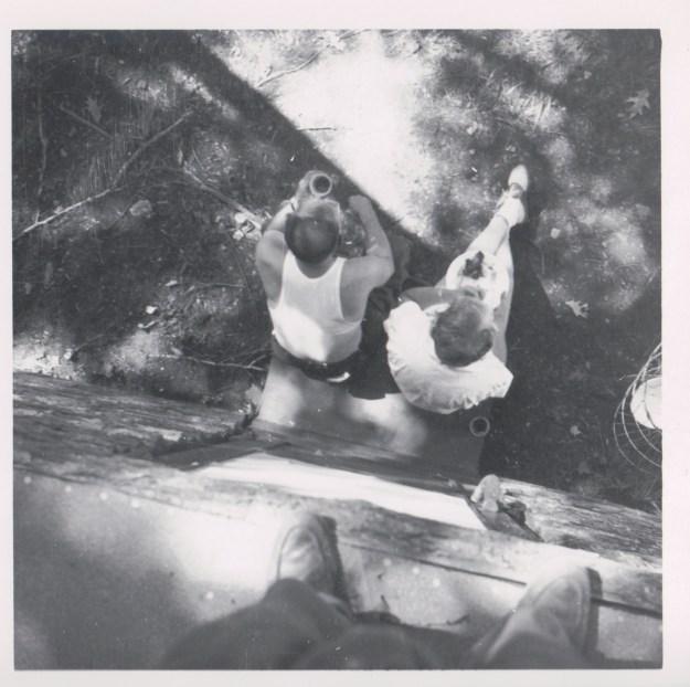 {Image:  black and white photo, taken from above looking straight down on two people sitting by the side of a stone building.  The person on the right is wearing saddle shoes and has their feet extended in front of them, with a plate of food on their lap.  The person on the left is wearing a sleeveless undershirt and holding a drink  (coffee?) in his left hand.  The photographer's feet on the edge of the building above are visible in the foreground.}