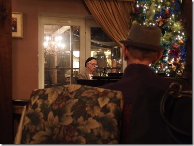 {Image:  photo.  in the left foreground, the back of a brocade (flowered) chair.  right foreground: the back of the head of an older gentleman with a short haircut and a hat.  To the right in the background, a Christmas tree with lights.  In the center, an older man in a beret playing the piano.}