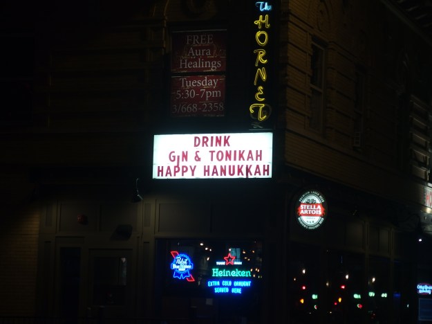 {Image:  lighted sign on the side of a bar reading "Drink Gin & Tonikah/Happy Hanukkah."  Above that sign is a vertical sign showing that the bar is The Hornet.}