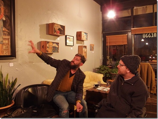 {Image:  Interior of a coffee shop with table and chairs (some wooden; some upholstered).  In the left side of the photograph, a man (same man as earlier photo - white man with short hair and beard) sits on the arm of a chair looking up toward a painting on the wall. One arm is extended toward the painting as he explains it.  To the left side of the photo is another white man, with a knit cap, glasses and a beard.  He also looks at the painting as he listens.}
