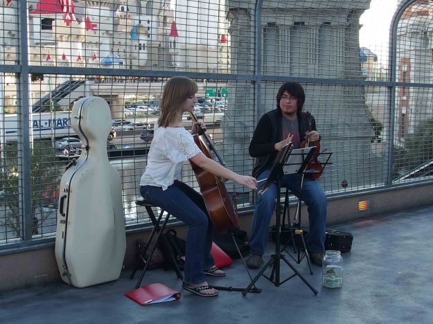 {A female cellist with cello and a male violinist with violin on a pedestrian bridge over a street in Las Vegas; the background full of neon signs and traffic.}