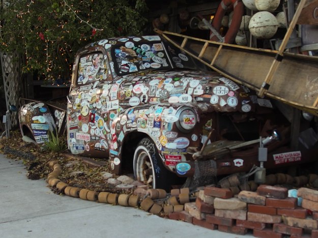 Image:   Old pick-up truck body, embedded in a brick sidewalk, covered with stickers.