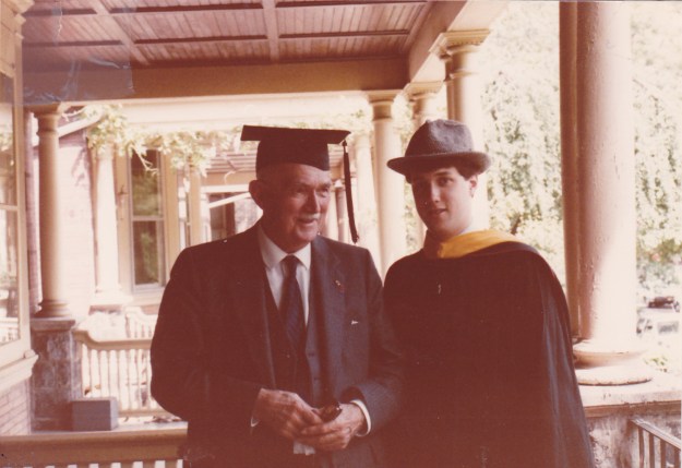 Image:  An older white man in a suit and tie who is wearing a graduation mortar board.  Standing next to him is a younger white man in a graduation cap and gown wearing the older man's fedora.  