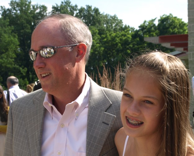Image:  white man in his 40s wearing sunglasses, a sports coat and an open-collar shirt; next to him is a white girl with long light brown hair.  