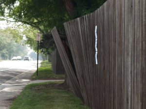 Image: a street runs along the left side of the photo; on the right  side, a green area bordered by a wooden fence which is sagging out into the green area next to the street. A white sign is visible tacked to the fence near the broken area. 