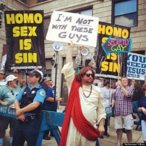 Image:  Protesters holding signs reading "Homo Sex is Sin."  They are behind a police line. In the foreground, a white man dressed as Jesus Christ holds a sign saying "I'm not with these guys."  Another counterprotester holds a sign pointing to one of the protesters that says, "Secretly Gay."  