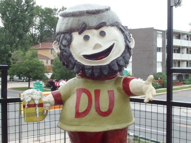 Image: larger-than-life-sized figure of a "pioneer" a white guy with a beard and a DU sweatshirt, holding a stein of beer in one hand.  His arms are open to the roof deck and he is smiling.  