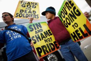Image:  Protesters (a white male adult and possibly light-skinned black child) holding signs that say "Thank God for Dead Soldiers," "Sin and Shame not Pride" and "You're Going to Hell."