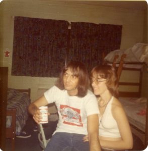 Image:  white man, 20-30 years old, wearing a Budweiser t-shirt and jeans, and holding a can of Budweiser beer shares a chair with a young white woman in a white halter top shirt.