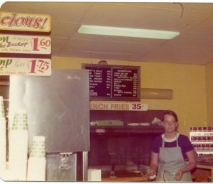 Image:  snack bar with white woman in apron behind the counter.  Sign reads French Fries 35 cents.