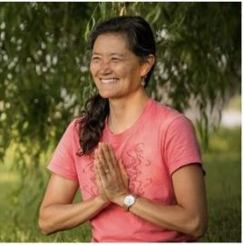 Image: Asian woman wearing rose colored t-shirt smiling with hands together in front of her in "namaste" gesture. 