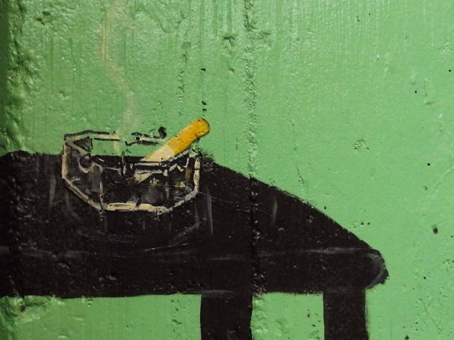 Photo of detail from a painted outdoor wall showing an ashtry with a half-smoked cigarette.