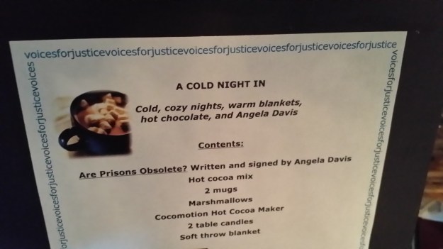 Image:  photo of a sign reading, "A cold night in:  Cold, cozy nights, warm blankets, hot chocolate, and Angela Davis.  Contents:  "Are Prisons Obsolete?" written and signed by Angela Davis; hot cocoa mix; 2 mugs; marshmallows; cocomotion hot cocoa maker; 2 table candles; soft throw blanket. 