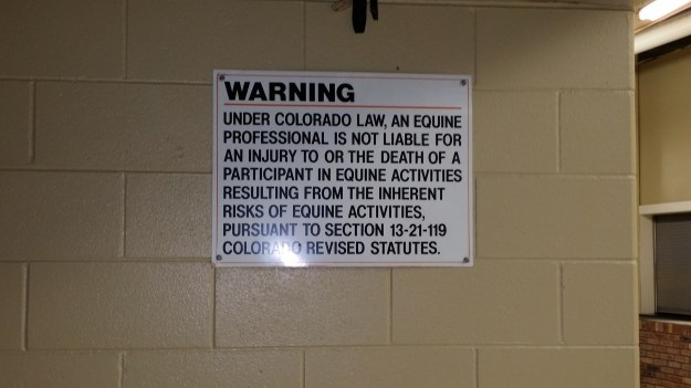 Image:  Sign affixed to a cinderblock wall that reads, "WARNING  Under Colorado law, an equine professional is not liable for an injury to or the death of a participant in equine activities resulting from the inherent risks of equine activities pursuant to Section 13-21-119 Colorado Revised Statutes. "