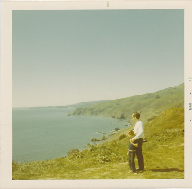 Photo:  A man and a girl (about 10) stand on the shore facing the water.  The man is in a white shirt and dark pants; the girl is in a blue shirt and pants.  The man has his hand on the girl's shoulder.