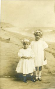 Image:  sepia photo of two young blond girls in white dresses with white ribbons in their hair, standing in front of a painting or backdrop of a beach with a rowboat. 