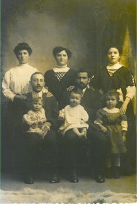 Image: sepia (brown and white) photo of eight people in formal dress of the early part of the 20th Century.  In the back row, three women (standing) in high-collared blouses, all apparently in their 20's or 30's; in the middle row, two men (sitting) wearing suits; the one on the left has a moustache; the one on the right has a full but neat beard.  In the front row, three children.  Two toddlers sit on the men's knees.  One perhaps 4-year-old stands on the right, with one of the women's hands on her shoulder.