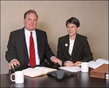 Tim and Amy at a conference table ca. 2000. (Tim is a white man with short blond hair who uses a wheelchair. He is dressed in a suit. Amy is a white woman with short brown hair and glasses. She is sitting in a chair, also wearing a suit. In the foreground, a table posed with law books, a speaker phone, files and mugs.