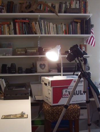 Image:  Camera set up to photograph objects on a table.  Lighting comes from a desk lamp on top of a cardboard box on top of a stool.  