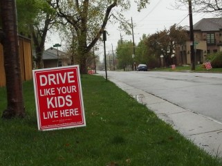 Image: Red lawn sign with white letters that read "Drive Like Your Kids Live Here."  Other lawns in the background have the same sign.