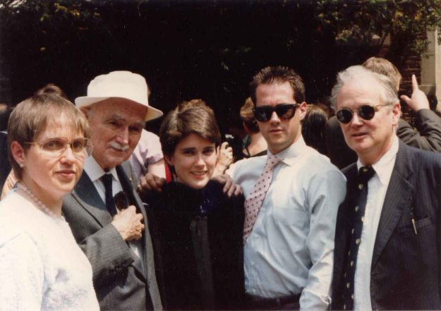 Image: 5 white people posing in front of a building. Mom (brown/gray hair and glasses in a white sweater); older man in suit and white fishing hat; me (short brown hair; graduation gown); younger man in spiked hair with sunglasses in a shirt and tie, and slightly older man in a suit and tie, also with sunglasses.