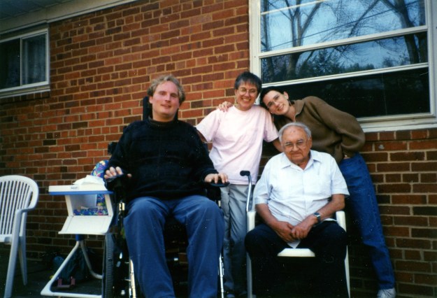 Image: four people, all white, posed on the patio in back of a brick house - man with blond hair in dark sweater using a wheelchair, woman in pink t-shirt, older (balding) man sitting in lawn chair wearing light blue shirt; me with an uncharacteristic pony tail wearing a brown sweatshirt and jeans. 