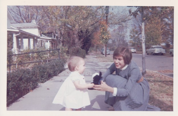 Image: White woman with brown hair in a blue dress squatting down to holdsa toy out to a white toddler in a white dress. They are on the sidewalk in front of a row of houses. 