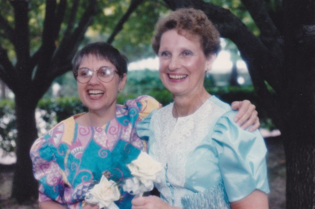 Image: two white women in party dresses, one with straight brown hair and glasses, the other with curly hair; both holding white roses.