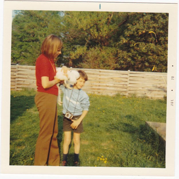 Image: white woman in shortsleeve red shirt holds a small white dog that is licking the face of a small boy in a blue sweatshirt and shorts with a camera around his neck.