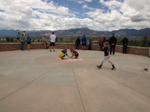 Image: people congregate on a concrete patio area facing the mountains while two young girls draw in chalk on the concrete and a young boy strides toward them.