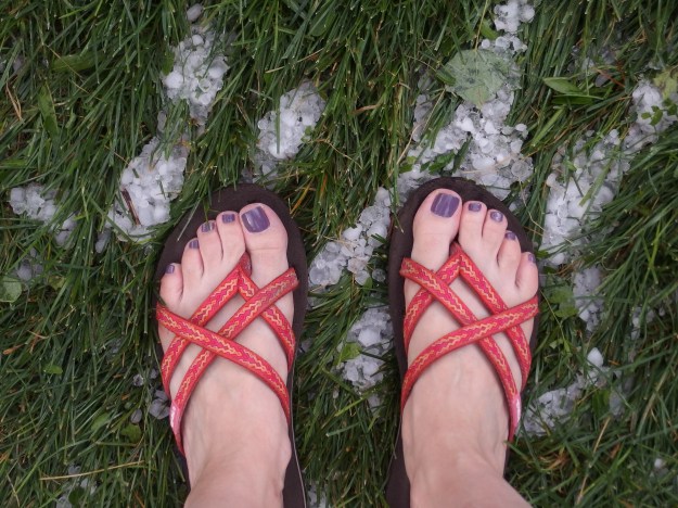 Image: photo of women's feet with purple toenails in flip flops standing on green grass covered with hailstones. 