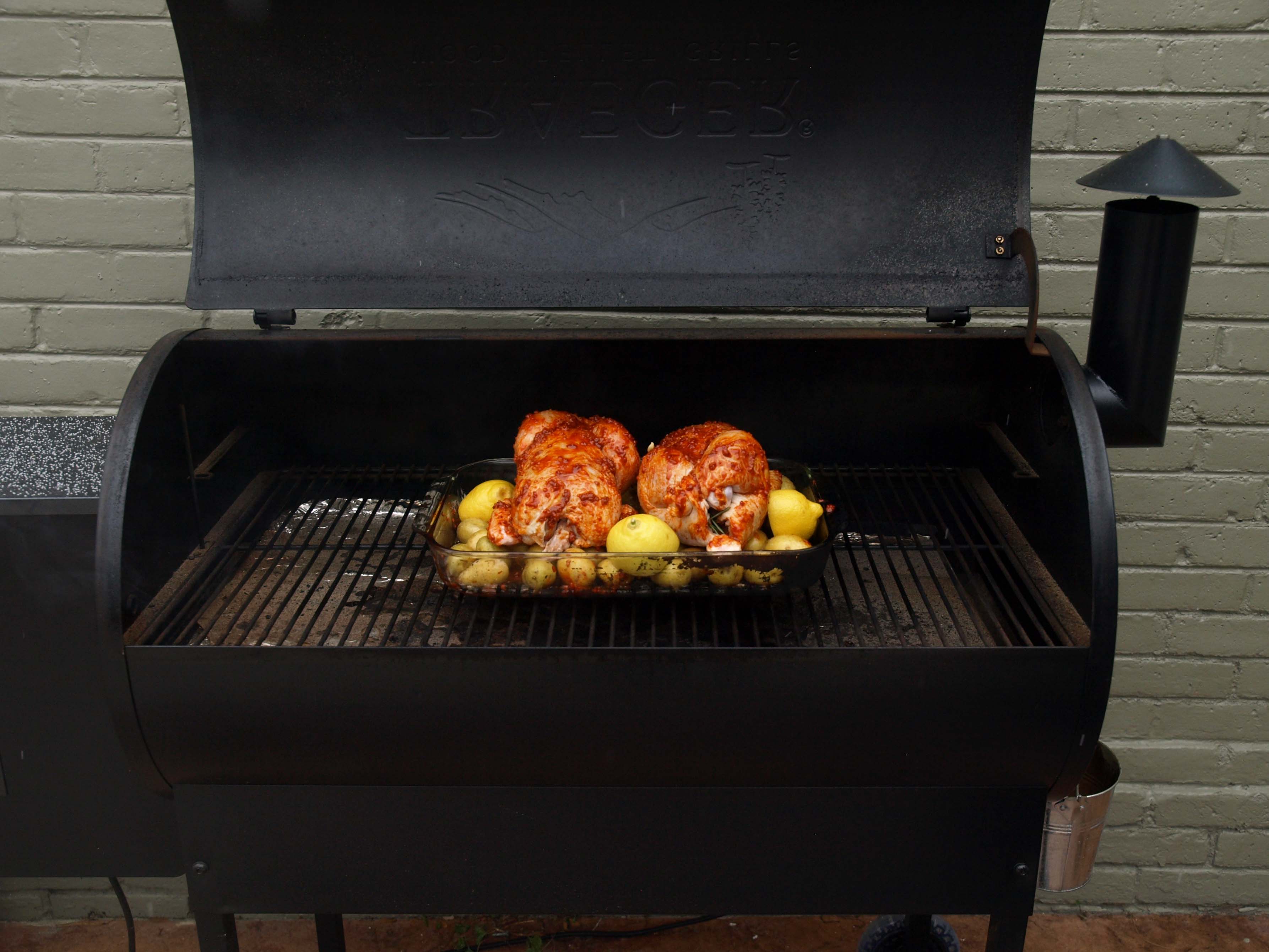 Image: Outdoor grill smoker with a glass baking dish containing potatoes and two whole chickens, spiced with smoked pimento.
