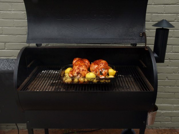 Image: Outdoor grill smoker with a glass baking dish containing potatoes and two whole chickens, spiced with smoked pimento.