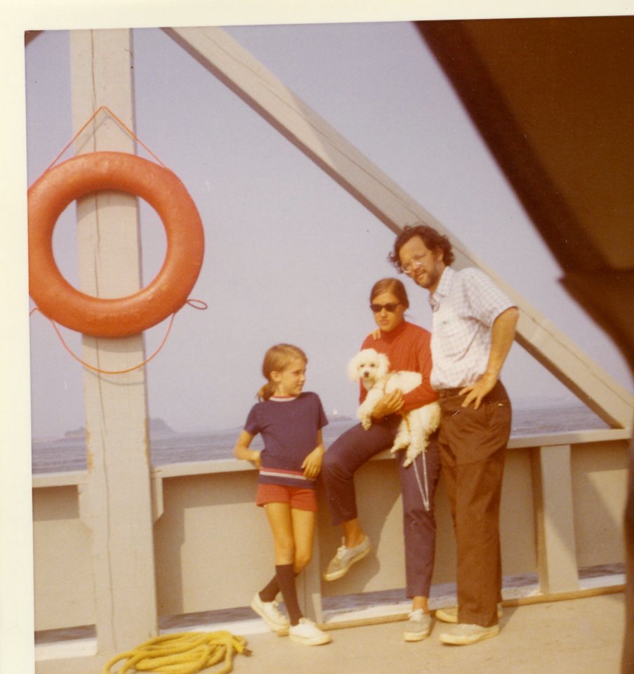 Image: three white people leaning on the side of a ferry boat. The young girl, around 9 years old, is wearing red shorts, a blue shirt and knee socks, the woman (holding a small dog) is wearing blue pants and a red shirt, and the man (with a scruffy beard) is wearing a short-sleeve button-down shirt and brown work pants.