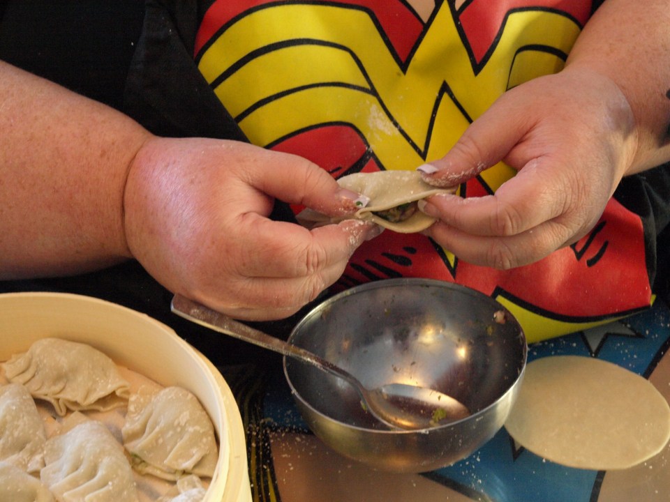 Image: close up of a pair of hands making dumplings next to a silver bowl of stuffing and a bamboo steamer of completed dumplings. Can see that the person is wearing a Wonder Woman logo shirt.