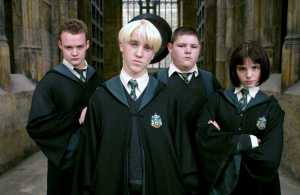 Image: four wizards from Harry Potter, middle school-age kids in black academic robes. All white. Second from left is Draco Malfoy, blond and sneering. To either side and slightly behind him are his sidekicks. 