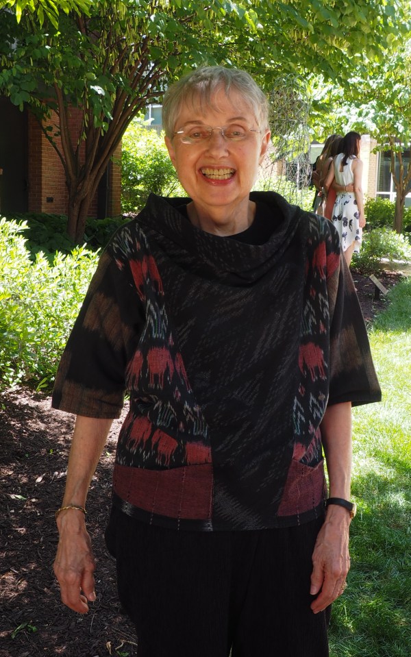 Image: Mom in 2016 standing in front of a leafy background, wearing a patterned black shirt and black pants.