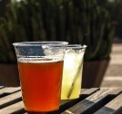Image: two drinks sitting on a wooden picnic table: a beer without a straw and a margarita with a straw.