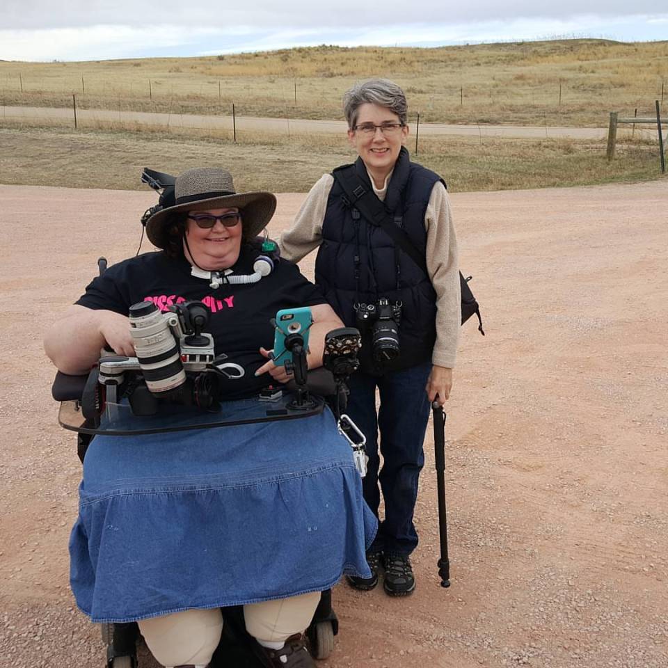 Two white-appearing women with cameras and other photo gear. Carrie, on the left, in a denim skirt and black t-shirt using a wheelchair. Amy, on the right, standing, wearing a beige fleece, vest, and jeans.