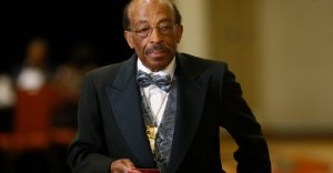Image: photo of Black man, balding with a mustache and wire-framed glasses, wearing a suit, a flowered bow tie, and a matching vest. 