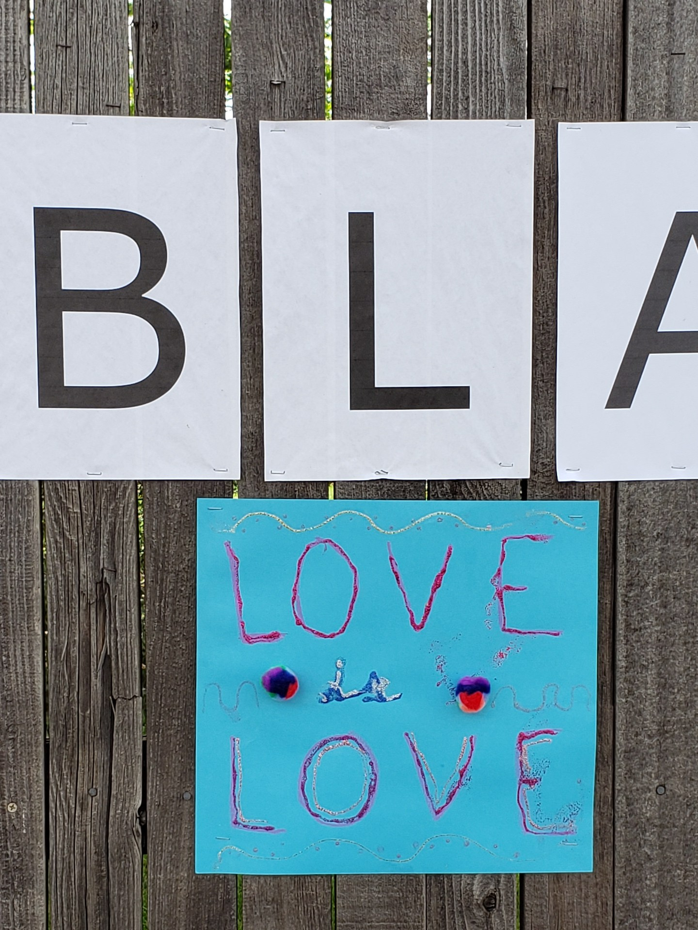 Blue construction paper sign that reads, "Love is Love" in red and blue ink with glitter and two multicolored cotton balls.