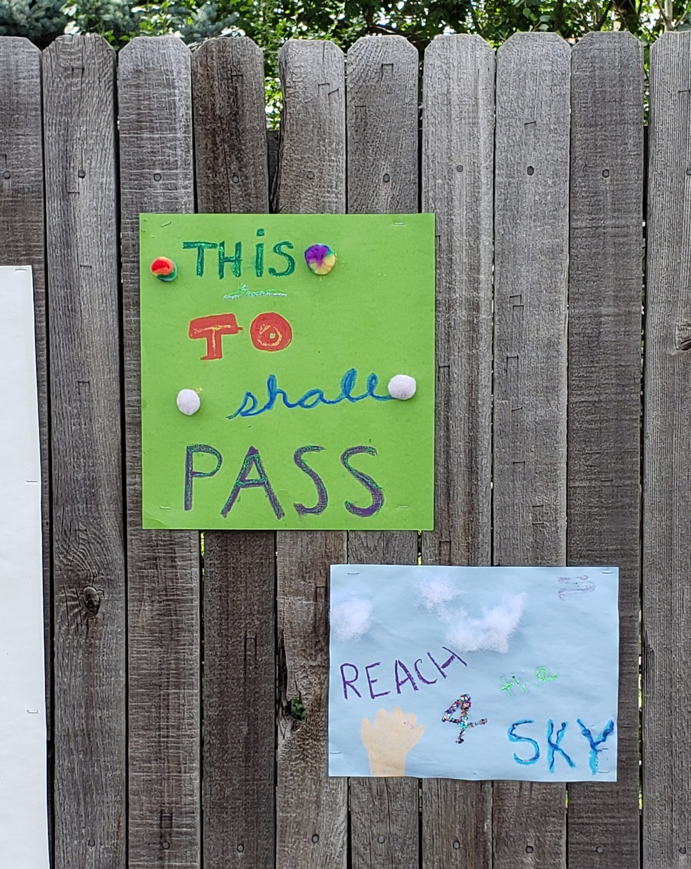 Two construction paper signs. One green that reads "This to shall pass," in various colors of ink with rainbow cotton balls dotted around. The second is on light blue paper and says Reach 4 the Sky with cotton ball clouds and a light-colored hand reaching up from below.